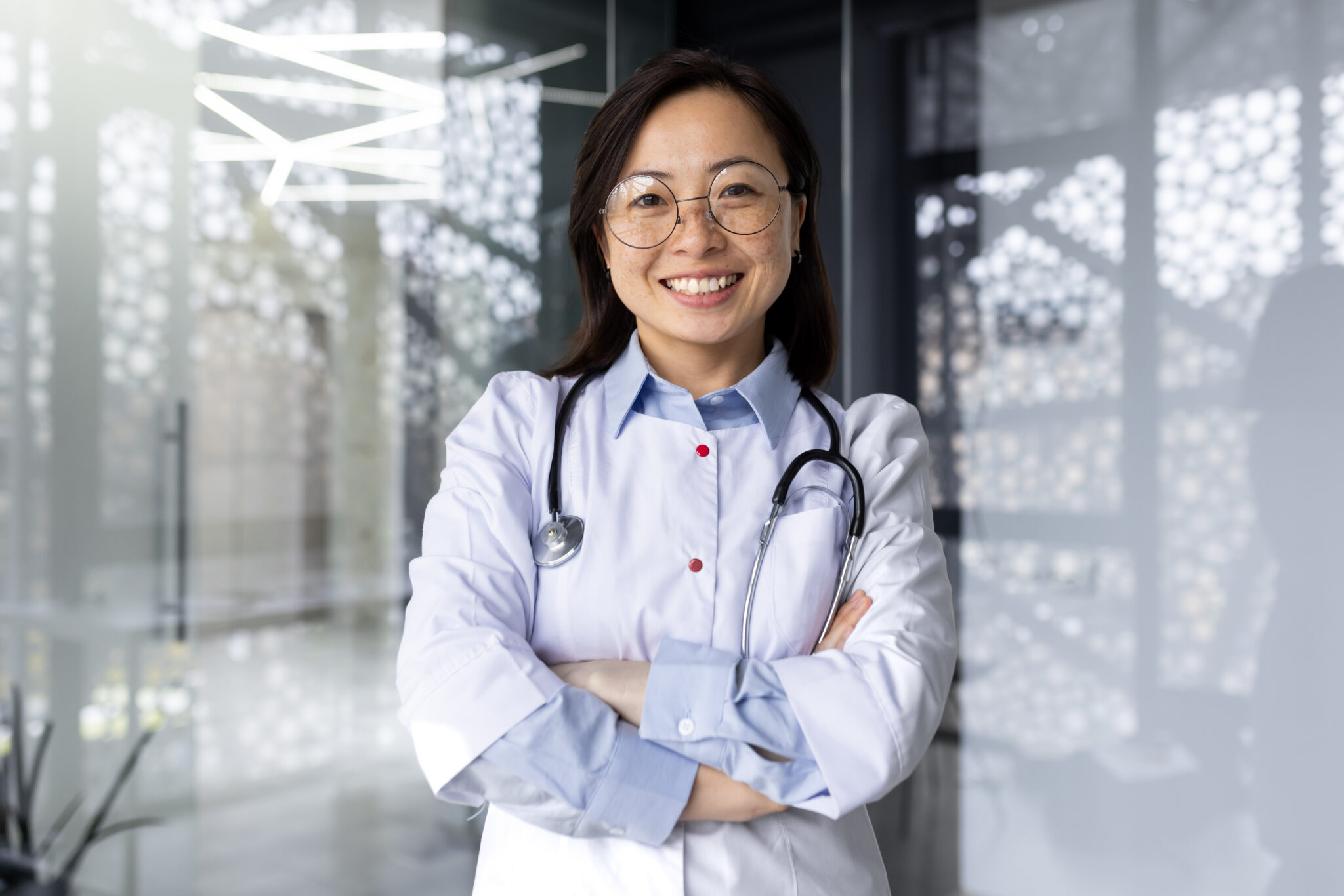 Portrait of a young beautiful and successful Asian woman inside a clinic office, a doctor smiling and looking at the camera with crossed arms and a white medical coat with a stethoscope